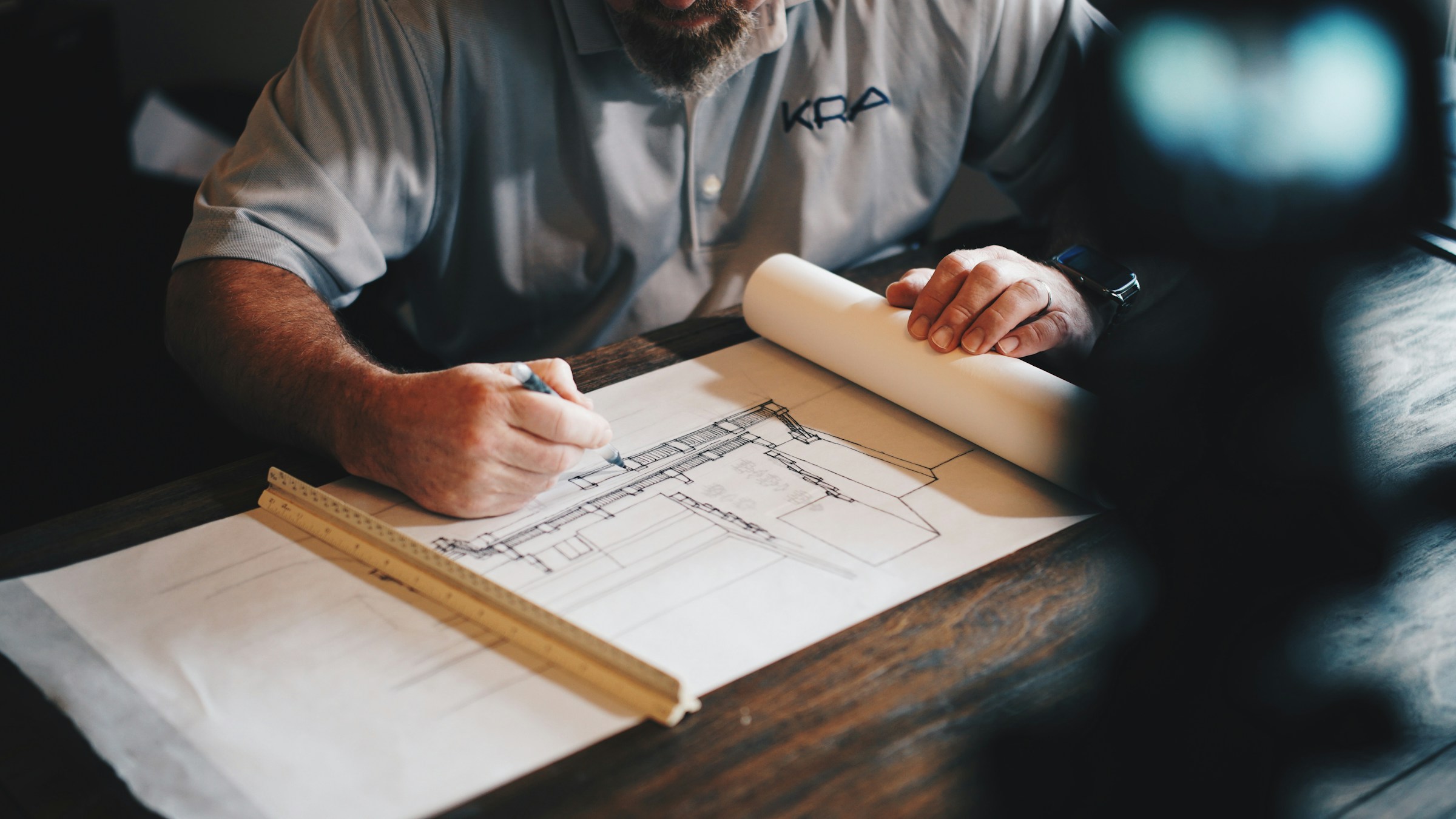 Hands marking a printed blueprint with a pencil and scale ruler on a wooden desk.