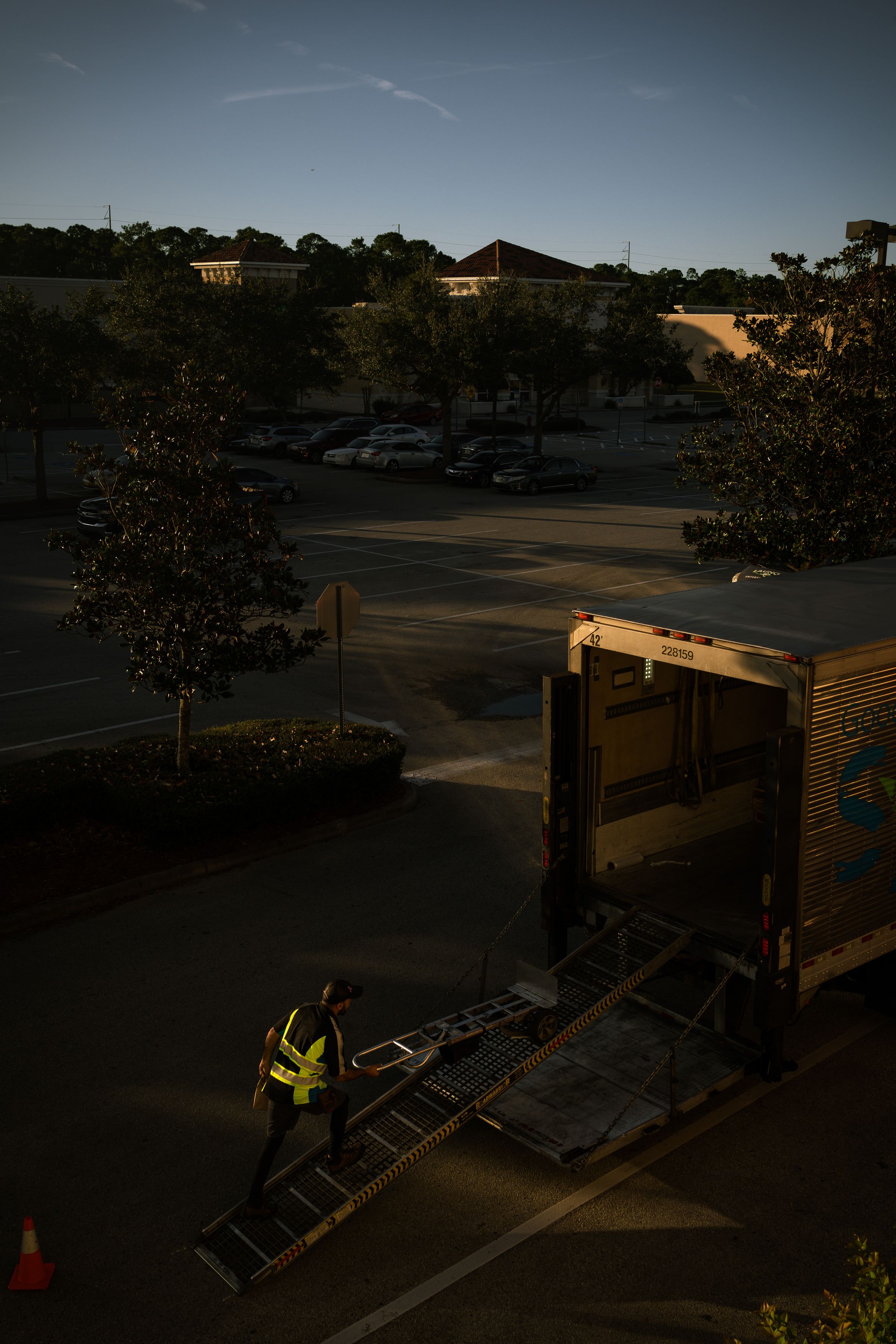 Worker loading a hand truck into the back of a box truck at a sunlit lot.