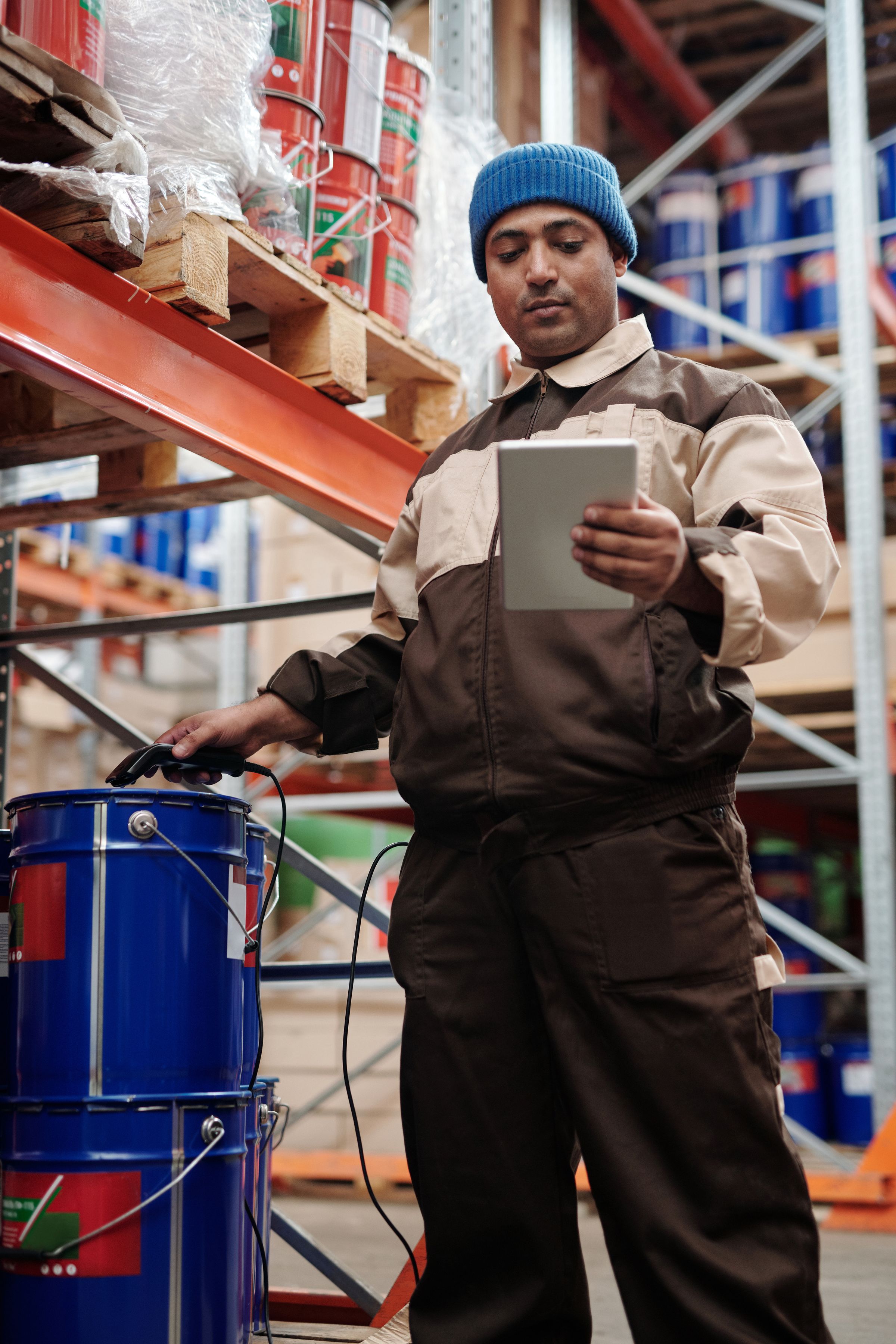 Warehouse worker in a work jacket checking stock on a tablet in an aisle of orange racking and paint pails.